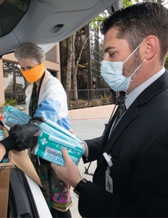 Two men in surgical masks load personal protective supplies into a car.