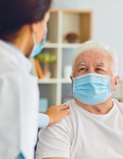 A masked female hospital staff member reaches out to comfort a masked older male patient.